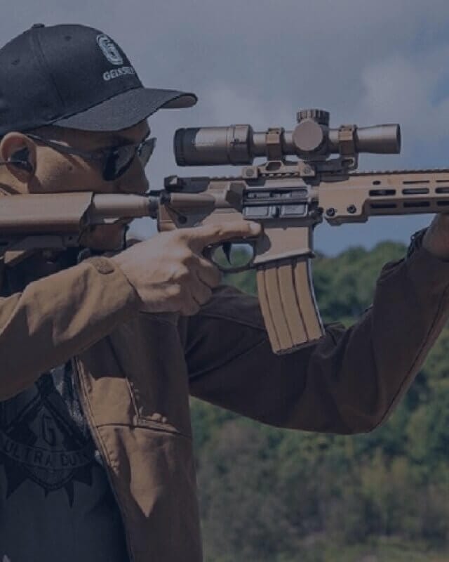 Geissele 67 Person aiming a scoped rifle at an outdoor shooting range while wearing protective eyewear and hearing protection.