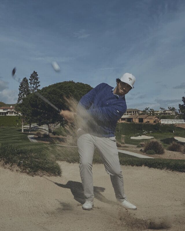 Golfer hitting a bunker shot during a round of golf, sending sand into the air while navigating a challenging course hazard.