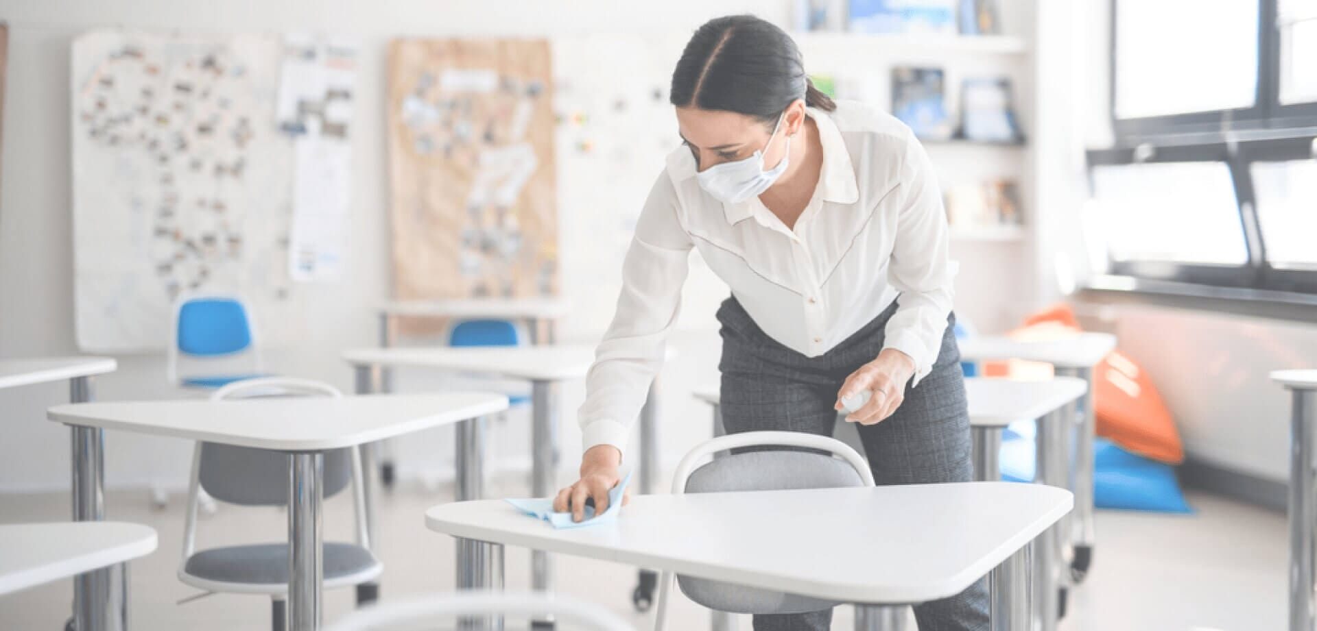 Professional cleaner wearing a face mask disinfects classroom desks in a bright educational setting, demonstrating commercial cleaning and sanitation services.