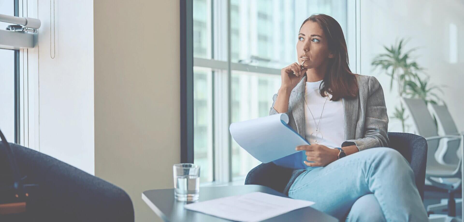 Professional woman reviewing documents in a modern office, seated near large windows while evaluating information and planning business strategy.