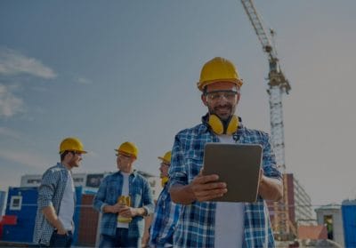 HVAC technician in a hard hat using a tablet on a job site, with crew members and construction equipment in the background.