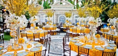 Elegant outdoor restaurant event setup with round dining tables, floral centerpieces, patterned plates, and bright yellow linens in a courtyard setting