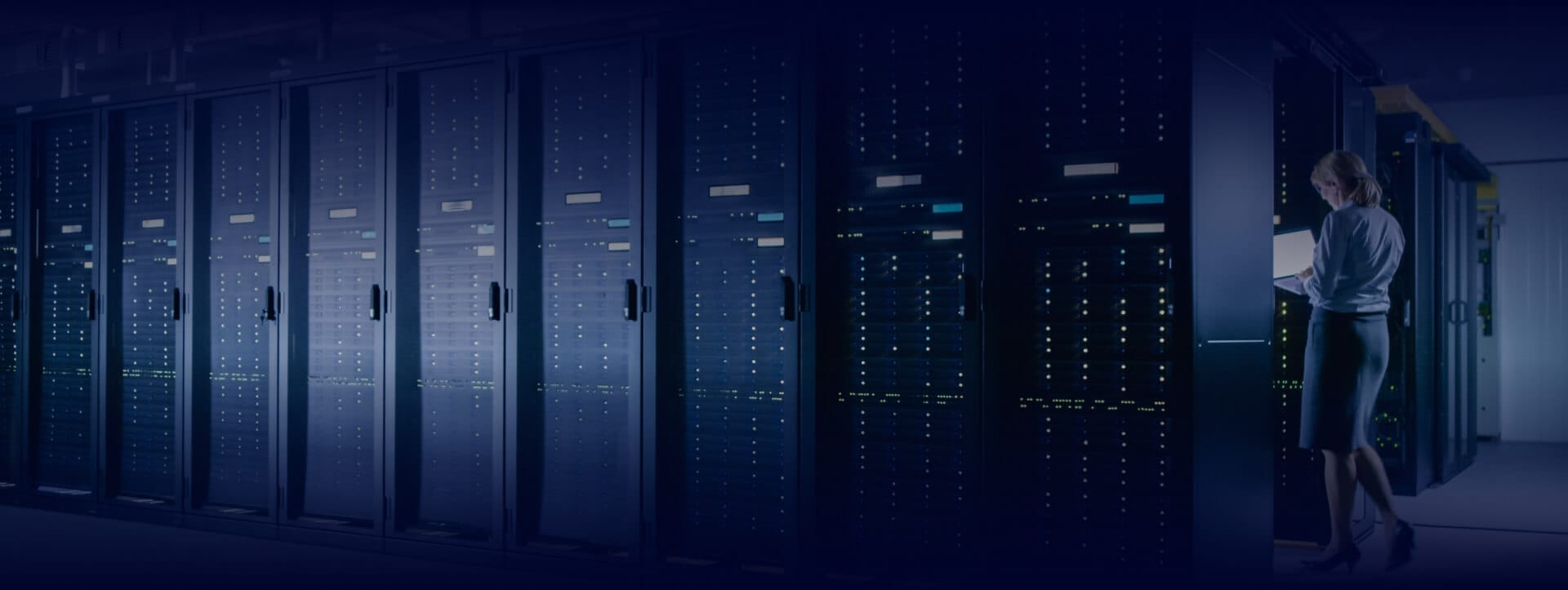 Woman reviewing a laptop in a dimly lit data center corridor lined with active server racks glowing blue