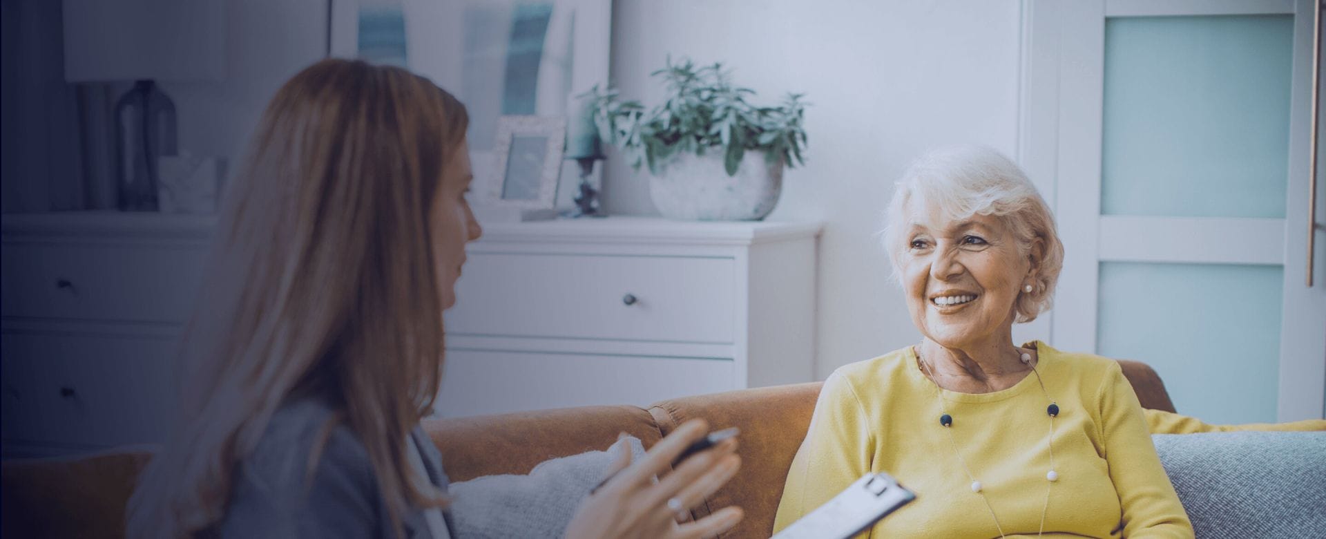 A healthcare professional speaks with a smiling older woman seated on a couch in a bright home setting