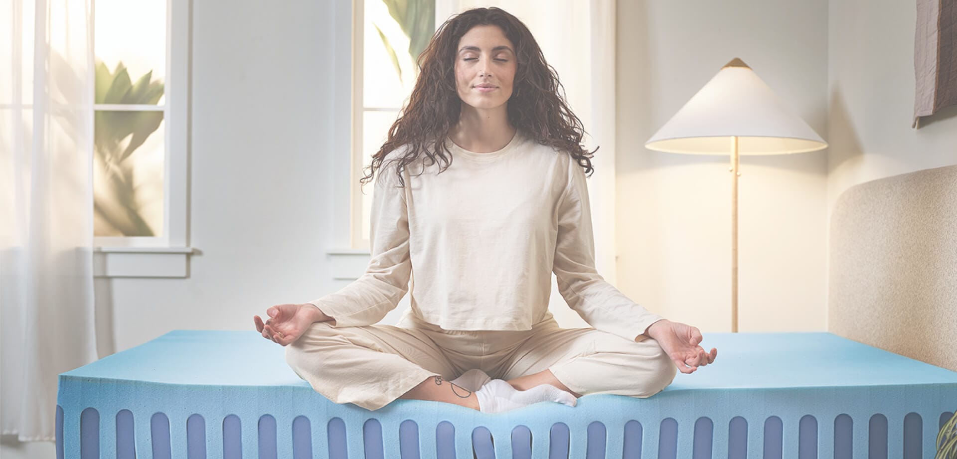A person sitting cross-legged on a blue mattress while meditating in a bright bedroom