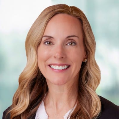 Smiling woman with wavy blonde hair wearing a dark blazer over a white top, photographed against a softly blurred light background