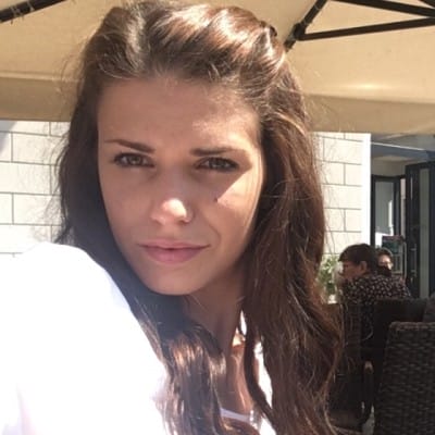 Close-up selfie of a woman with long brown hair, wearing a white top and looking at the camera while seated outdoors at a café in bright sunlight