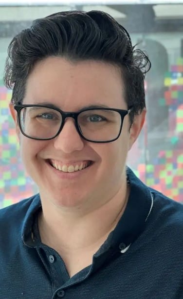 Home Page - NEW 247 Smiling person with dark hair and black glasses wearing a navy collared shirt, with a colorful blurred background behind them