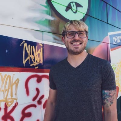 Web Design 81 Smiling man with glasses and a tattooed arm wearing a dark T-shirt, standing in front of a colorful graffiti-covered wall