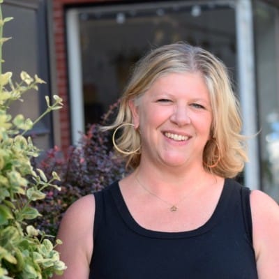 Brand Identity 71 Smiling woman with blonde hair wearing hoop earrings and a black sleeveless top, standing outdoors near plants and a storefront