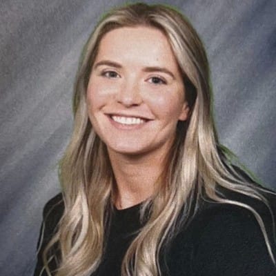 Smiling woman with long blonde hair wearing a black top, photographed against a gray studio-style background