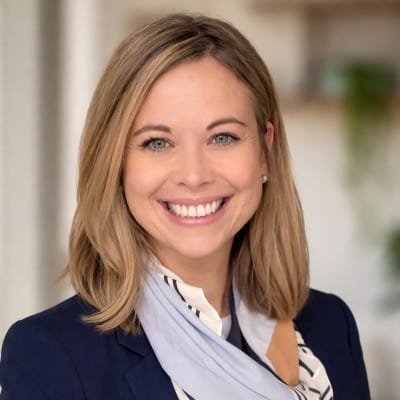 Professional headshot of a smiling woman with shoulder-length blonde hair, wearing a navy blazer and light scarf against a softly blurred indoor background