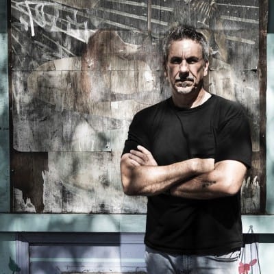 Man wearing a black T-shirt and jeans, standing with arms crossed in front of a textured wall with abstract artwork