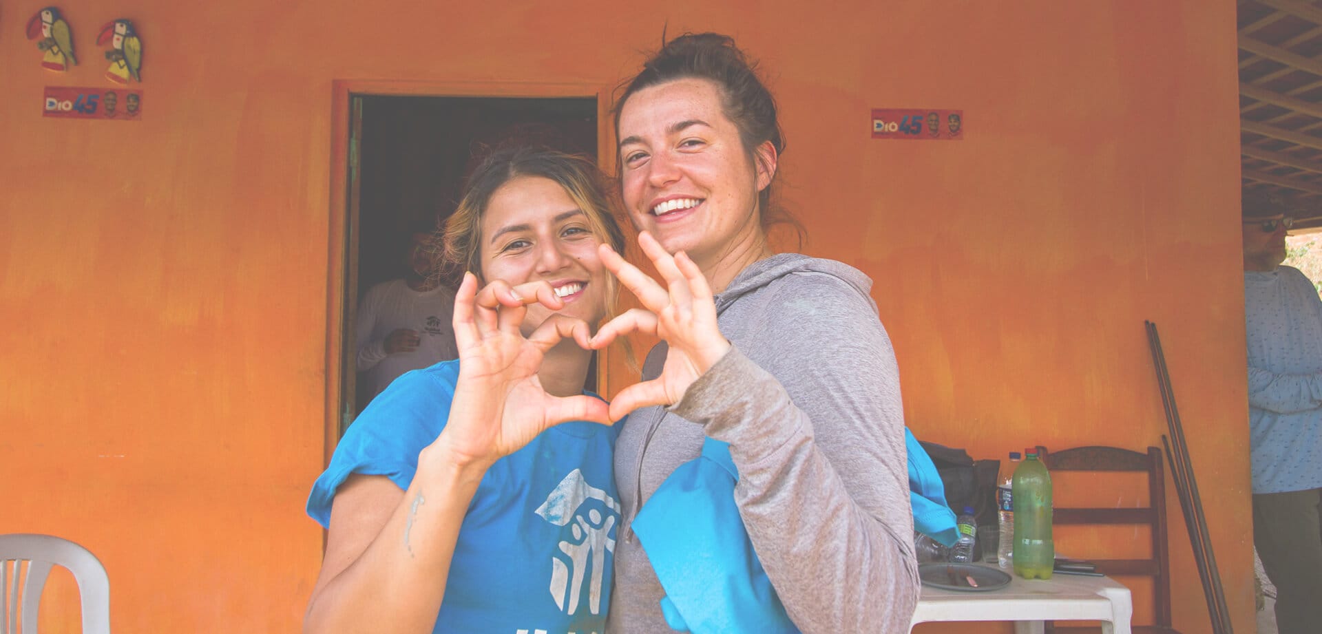 Two smiling volunteers stand in front of an orange wall, forming a heart shape with their hands while wearing blue Habitat for Humanity shirts