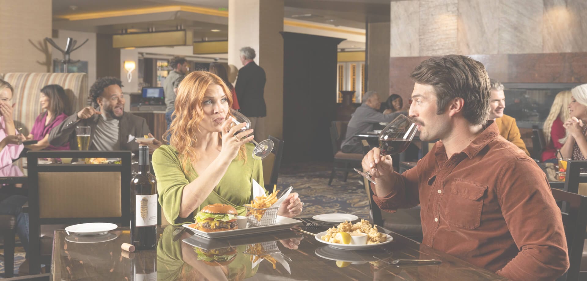 Couple dining in a busy restaurant, drinking wine with burgers and appetizers on the table