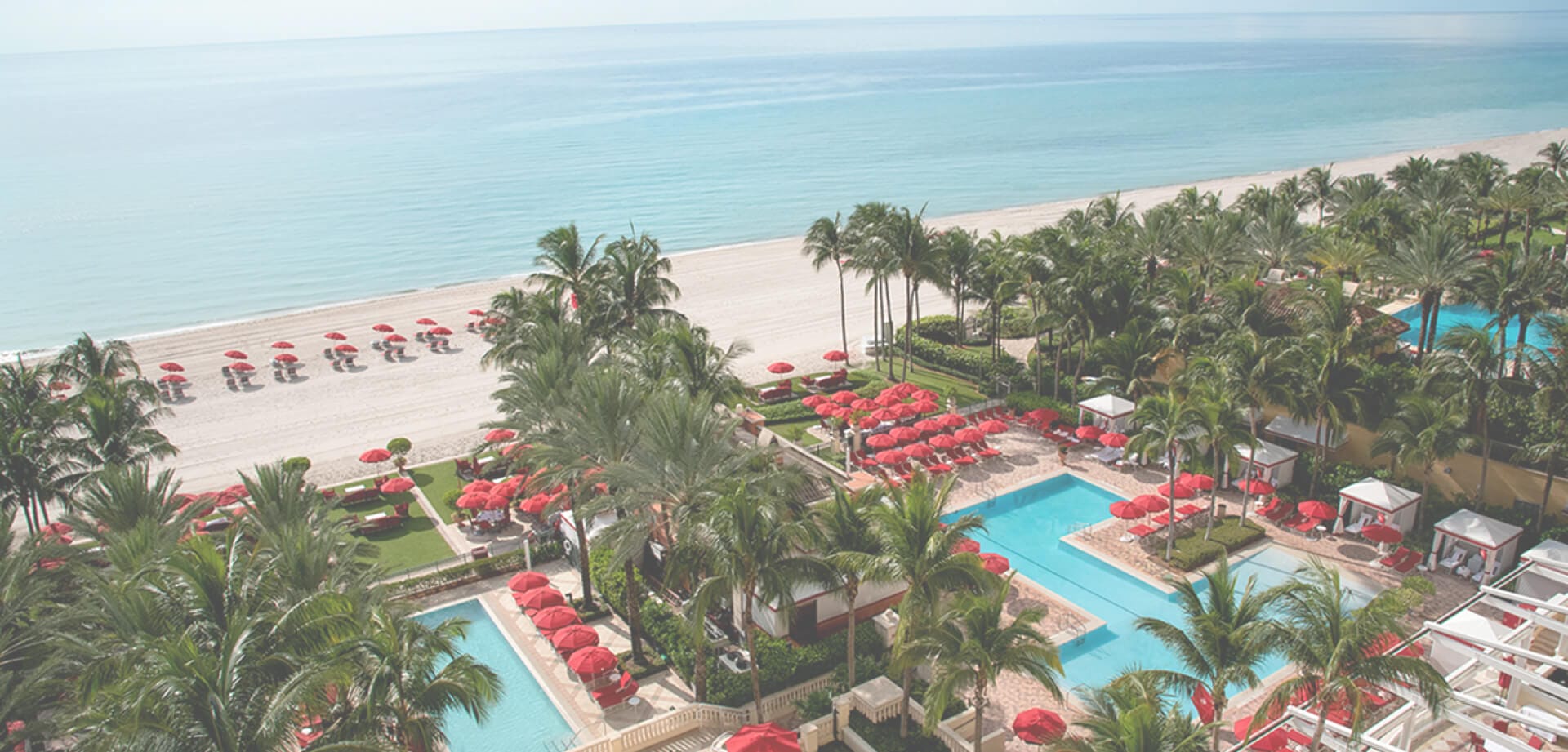 Aerial view of a beachfront resort with turquoise swimming pools, palm trees, red umbrellas and lounge chairs, beside a wide sandy beach and calm blue ocean
