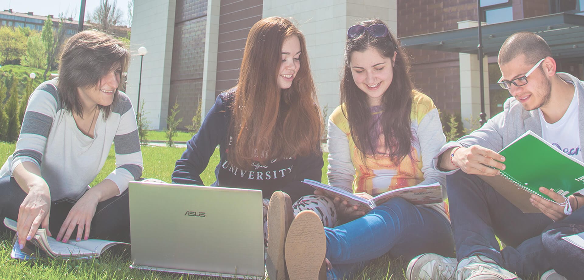 Four university students sit on the grass outside a campus building, smiling as they study together with books, notebooks, and a laptop